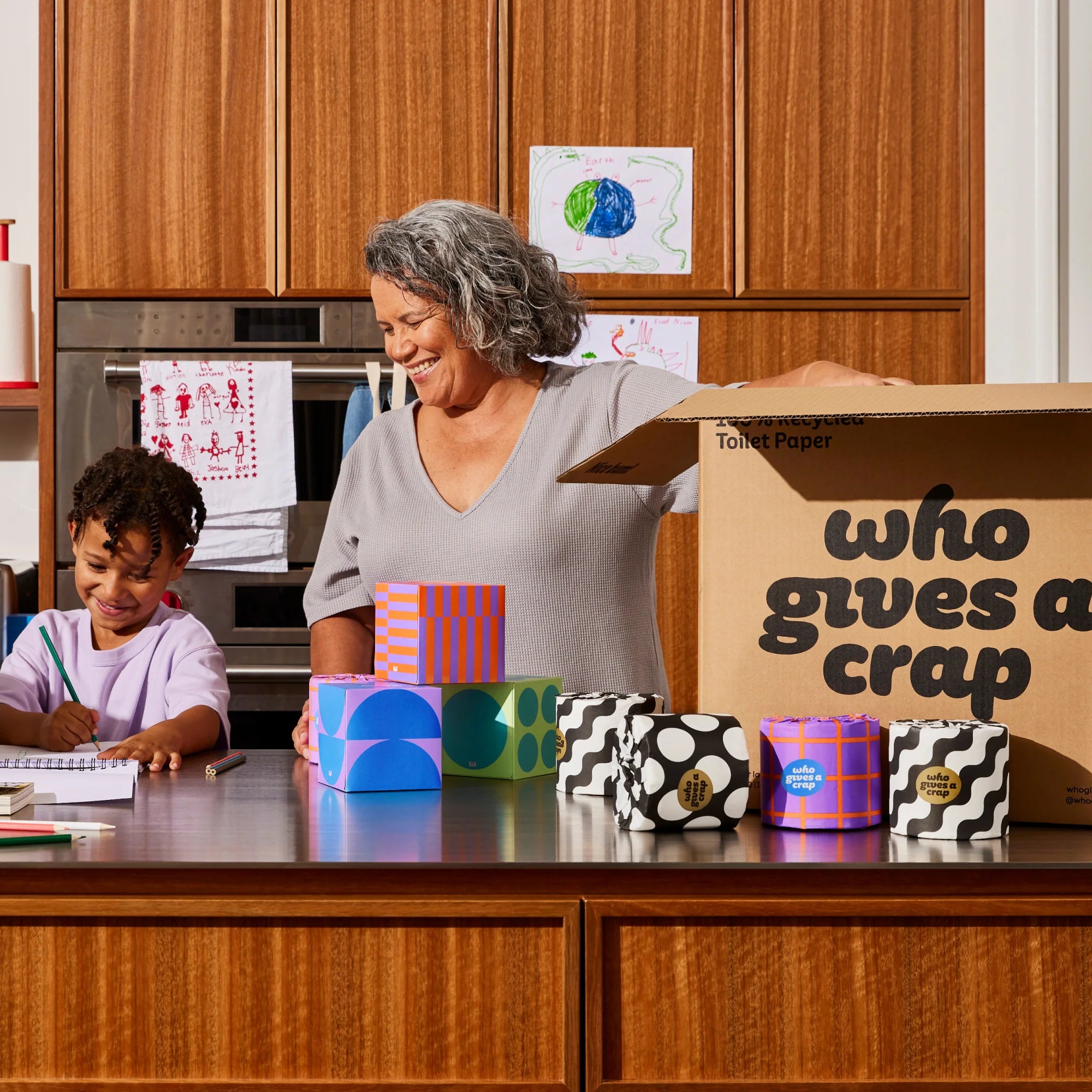Person unboxing Who Gives a Crap products onto the kitchen benchtop, while a young child is colouring on paper